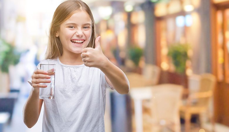 A girl giving the thumbs up while holding a glass of water.